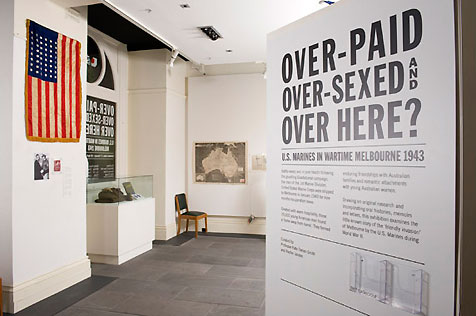 Gallery entrance to Over-Paid, Over-Sexed and Over Here, showing intro wall text on right, USA flag on left and Australian map in central background