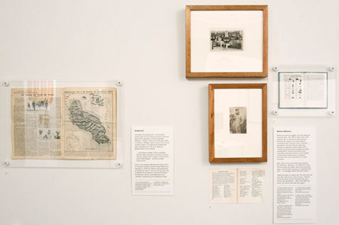 Two framed black-and-white photographs and five pieces of unframed ephemera hang on the wall, the piece on the left showing an island map