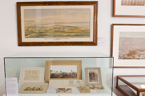 Display case with six sepia photos and other paper ephemera; three framed watercolours hang on wall behind