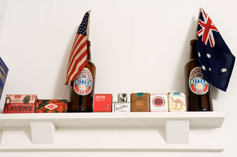 Detail of a shelf holding old cigarette packets and two beer bottles, one with an American flag on a stick, the other with an Australian flag