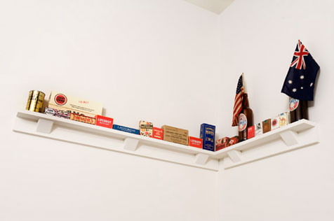Corner shelf holding old cigarette packets, product packaging and two beer bottles, one with a US flag on a stick, the other with an Australian flag