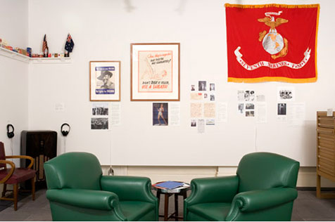 Gallery’s east wall, hung with ephemera and a large red banner at the upper right; two green armchairs are in the centre foreground