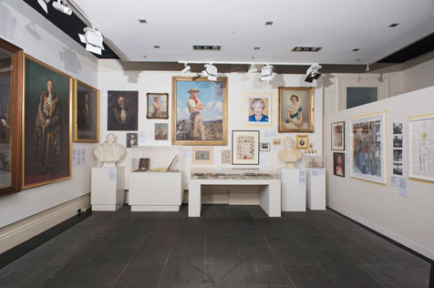 Front-on view of the interior of the gallery showing multiple portrait paintings on three walls, two busts on white display stands and a white display table
