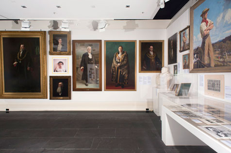 Corner of the gallery with 11 portraits on the walls, a glass display cabinet and white bust