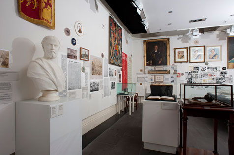 Main wall of gallery showing white bust, brightly coloured tapestries, photos and dark portrait, alongside other ephemera