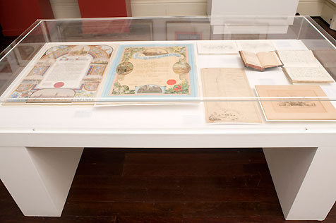 Glass-topped display case holding a book and paper ephemera; two decorative, colour certificates are on the left of the display case