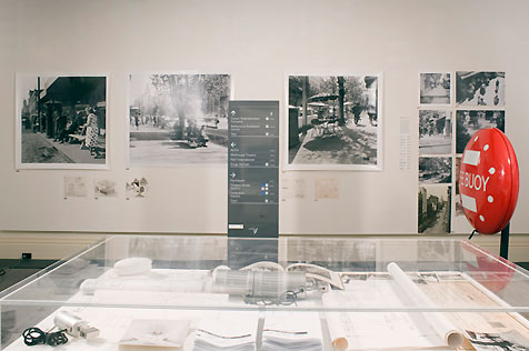 Gallery’s north wall, hung with three large black-and-white photographs and eight small; a glass display case in foreground holds paper ephemera