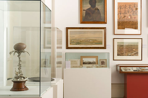 Exhibition display with emu egg on ornate silver stand; four paintings on wall and Aboriginal portrait