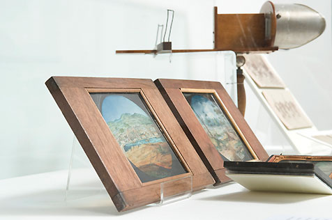 Timber-framed painted scenes in display case, with an old image viewer and sepia photographs in the background