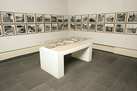 North-east corner of gallery, showing table display case and wall-mounted sequence of 32 framed black-and-white photos in two rows