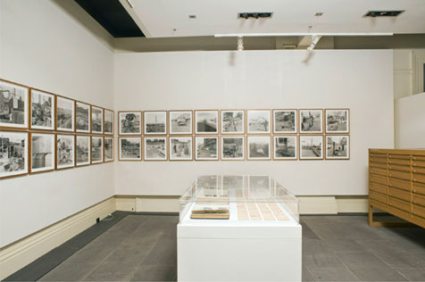 North and east walls of gallery hung with 30 framed black-and-white photos, table display case in foreground, wooden display cabinet on right
