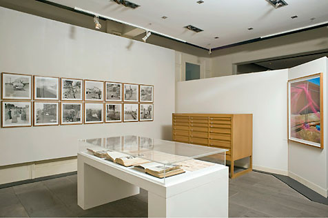 East and south walls hung with 14 black-and-white photos and large colour image, on right; table display case in foreground, wooden cabinet behind
