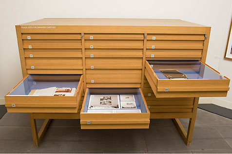 Wooden display cabinet with three of its 21 drawers open, displaying printed material in exhibition