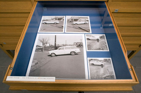 Open drawer of display cabinet showing five black-and-white photos, one large four small, of a car crossing a bluestone on a road intersection