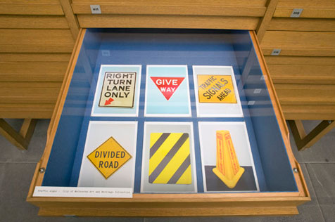 Open drawer of wooden display cabinet showing six colour photos of road signage; five are flat signs, one a yellow ‘witches’ hat’