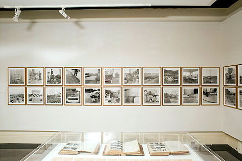 North wall of gallery hung with 24 framed black-and-white photos in two rows; a table display case holding three open ledgers is in foreground
