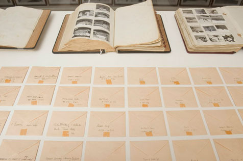 Detail of display case, with grid of 30 square envelopes in foreground and three open ledgers behind, in which 19 black-and-white photos appear