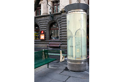Outside the gallery, with green street seating at left and technical drawings of street lighting in a tall cylindrical display case on the right