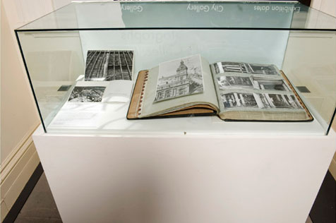 Glass-topped display case with open ledger showing six mounted black-and-white photos; two black-and-white photos are to left of ledger