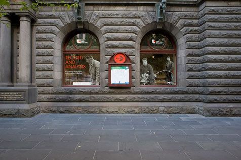 Gallery’s two arched windows in the Town Hall’s bluestone façade, with advertising for Record and Analysis on windows; pavement in foreground