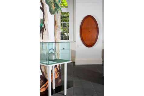 Exhibition installation including a kettle in a glass case and oval wooden table top hanging on a wall