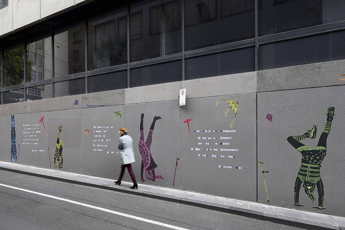 A person walks along a laneway. There are stencilled images on the walls, showing people doing handstands with a walking aide nearby.