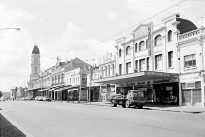 A black and white photograph of a North Melbourne street in the 1960s. Cars are parked outside of businesses.