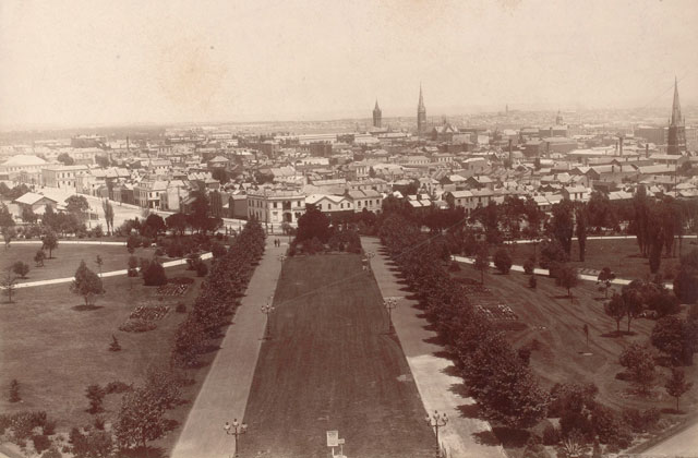 Black and white photo showing an elevated view over a wide tree-lined avenue, leading to city buildings in the background.