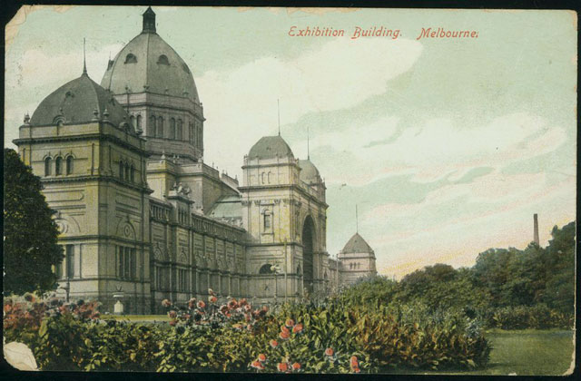 Re-coloured black and white photograph showing the Exhibition building with flowering plants in the foreground.