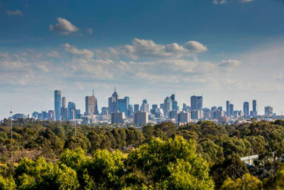 Melbourne CBD buildings under a blue sky, with trees and bushland in the foreground.