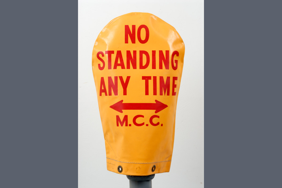 Yellow parking meter cover with the words in red: 'No standing any time M.C.C' and arrow pointing left and right