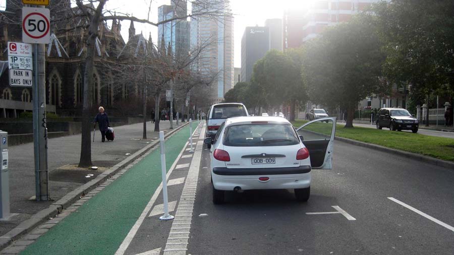 The Albert Street bike lane with the kerb on the left and parked cars to the right. It is separated from the parked cars by wide striped line-markings, rumble strips and bollards.