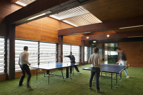 Four men playing table tennis on two tables in a room with louvered windows and fake grass on the floor.