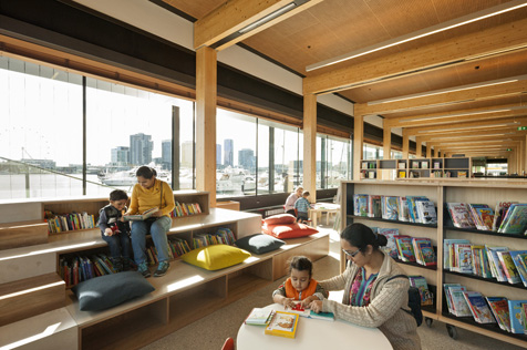 Women and children sitting reading books in a room with bookshelves and windows on to a harbour with moored boats and high rise buildings in the background.