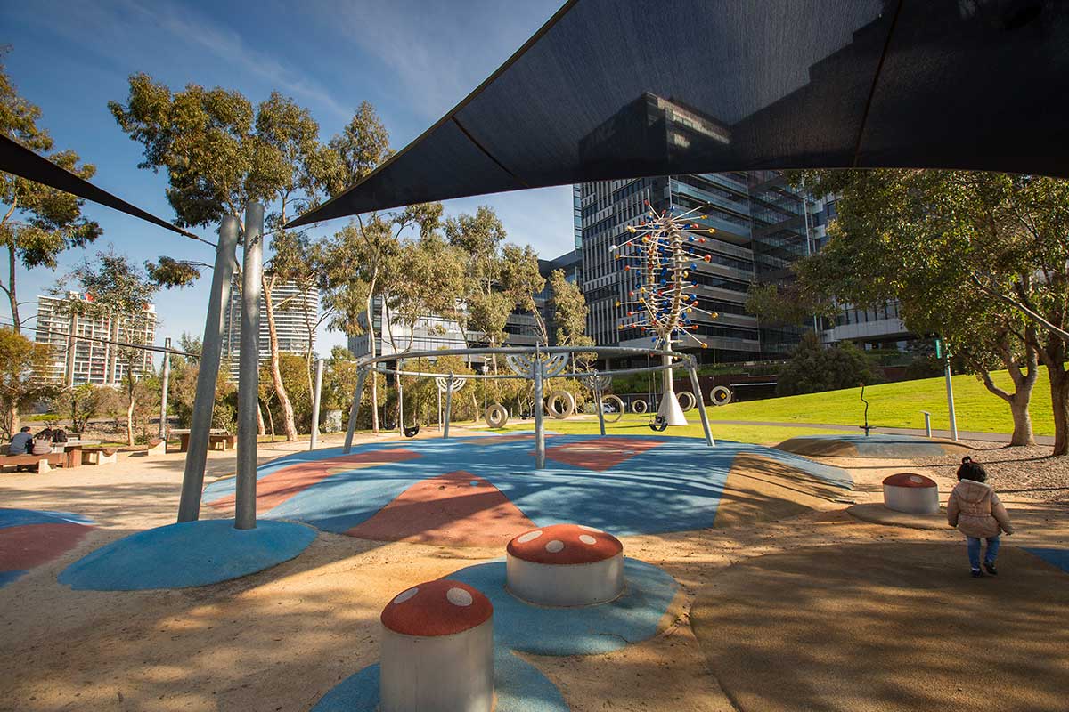 Playground in Docklands Park with climbing platforms decorated like mushrooms, swings and other play equipment, under a shadecloth.