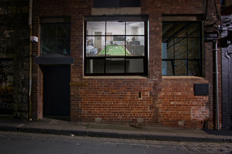 Red brick building facade with image of an interior containing a billiard table and other furniture projected on its window