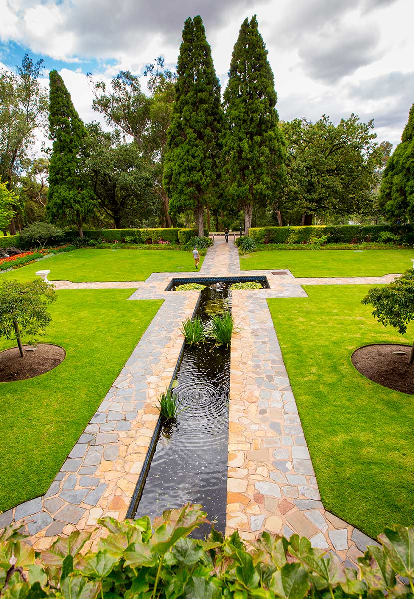 A long, narrow pool with symmetrical paved edging, grass and formal trees planted alongside.