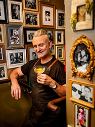 Alan Bell holding a cocktail in Cabinet Bar. The surrounding walls are decorated with framed black and white photos.