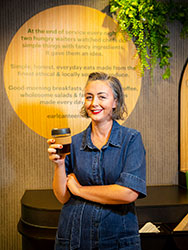 Jackie Middleton with coffee in a reusable cup, standing in front of a circular sign on a wall at EARL Canteen.