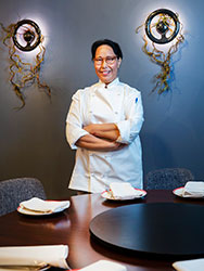 Jagjeet Kaur in chef's whites standing in front of designer wall-mounted lights amd next to a round dining table set formally with folded napkins, plates and a lazy susan.