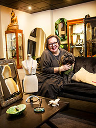 Kirsten Albrecht sitting on a sofa with a dog, surrounded by decorative objects, jewellery, glass display cabinets and mirrors. 