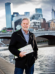 Tim Bracher standing next to the Yarra River, with a bridge and city buildings in the background.