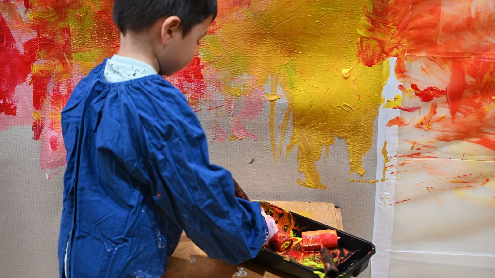 Child using a roller to paint on a canvas in yellow and red.