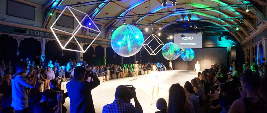 Fashion show in the Meat Market space with large cube and sphere decorations suspended from the ceiling. Models are walking in the central runway space, surrounded by an audience and photographers crowded around the edges of the room.
