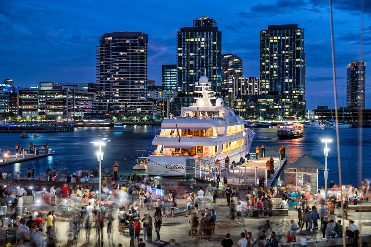 A yacht moored at Melbourne City Marina at dusk, with crowds of people socialising on the waterfront