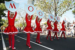 Row of girls dressed in red, holding up letters spelling out 'Moomba' in parade