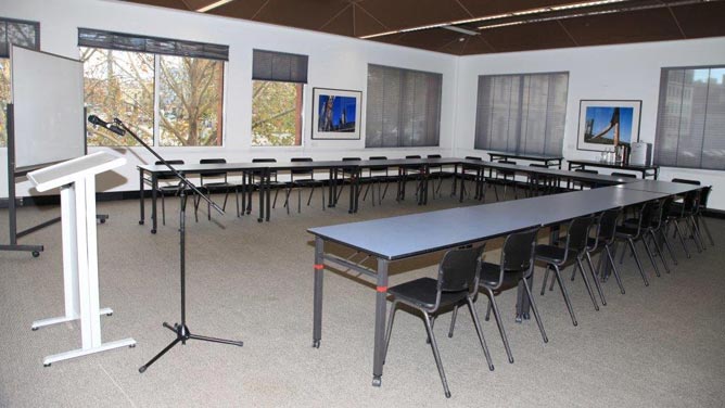 Room with tables and chairs arranged in U-shape, facing a lectern with microphone and whiteboard. There are windows on two sides