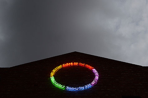 Circular neon sign on gabled building, in spectrum of green, yellow, orange, red, purple and blue spelling out the words 'to the point where we can see nothing everything has become clear'