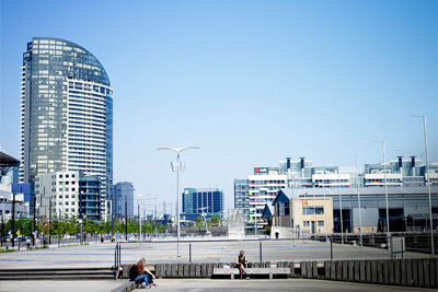 A photograph of people sitting on a boardwalk in Docklands. Melbourne skyscrapers are in the background. 