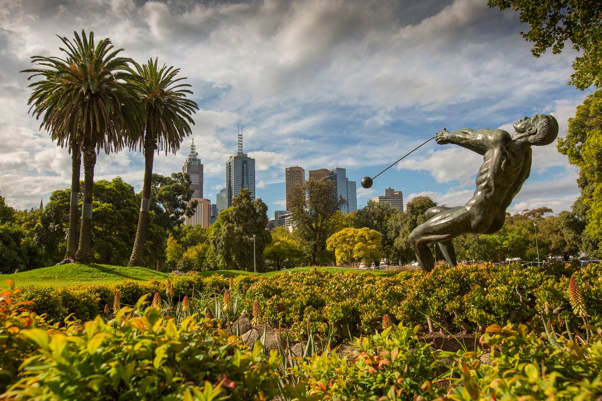 Bronze sculpture of a naked hammer thrower in action, leaning backwards. The scupture is set among garden beds with palm trees nearby and CBD buildings in the background.